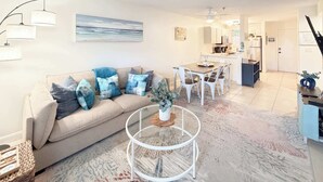 A modern living room with a beige sofa, a glass coffee table, and a dining area with white chairs. - Coastal 2 bed 2 ba Villa - Pools - beach access (Hilton Head Island)