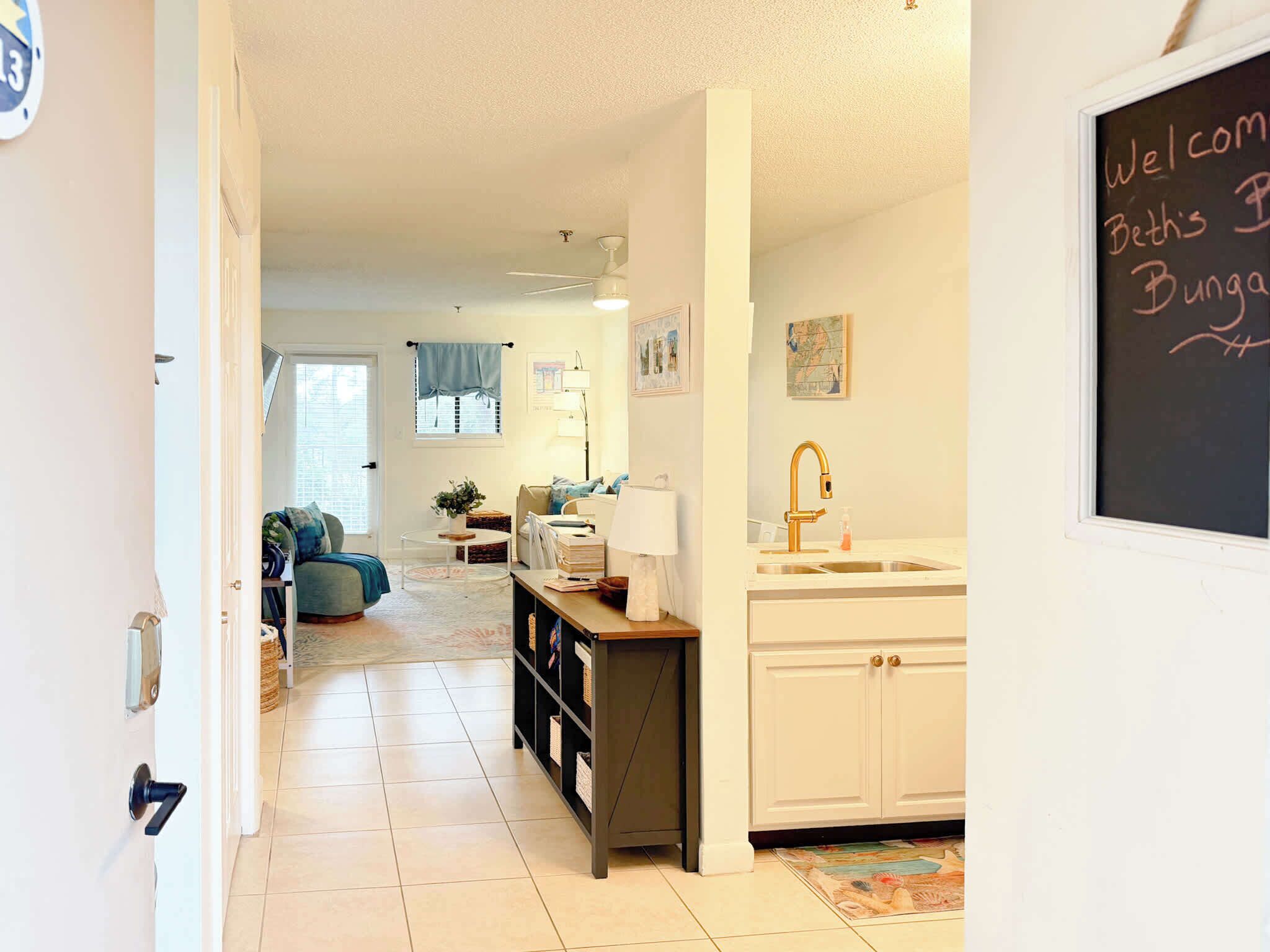 A cozy living room with a sofa, a coffee table, and a bookshelf. There is a kitchen area with a sink and cabinets. A chalkboard with "Welcome" and "Beth's Bungalow" is visible.
