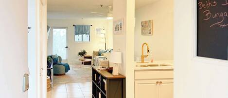 A cozy living room with a sofa, a coffee table, and a bookshelf. There is a kitchen area with a sink and cabinets. A chalkboard with "Welcome" and "Beth's Bungalow" is visible.