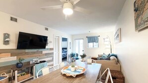 A modern living room with a wooden dining table, white chairs, a flat-screen TV, and a cozy seating area with a sofa and cushions. - Coastal 2 bed 2 ba Villa - Pools - beach access (Hilton Head Island)