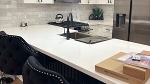 A modern kitchen with white cabinetry, a stainless steel refrigerator, a microwave, and a gas stove. There are two black upholstered bar stools and a box on the counter. - High End Luxury 3bedroom Located Near Downtown Houston (Houston)