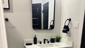A bathroom with a white sink, black soap dispenser, and a mirror above it. There is a black trash bin and a toilet paper roll on the wall. A black bag hangs on the wall next to a light switch. - High End Luxury 3bedroom Located Near Downtown Houston (Houston)