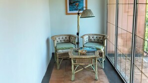 Een kamer met twee rieten stoelen, een tafel, een vloerlamp en een groot raam met uitzicht op de omgeving.