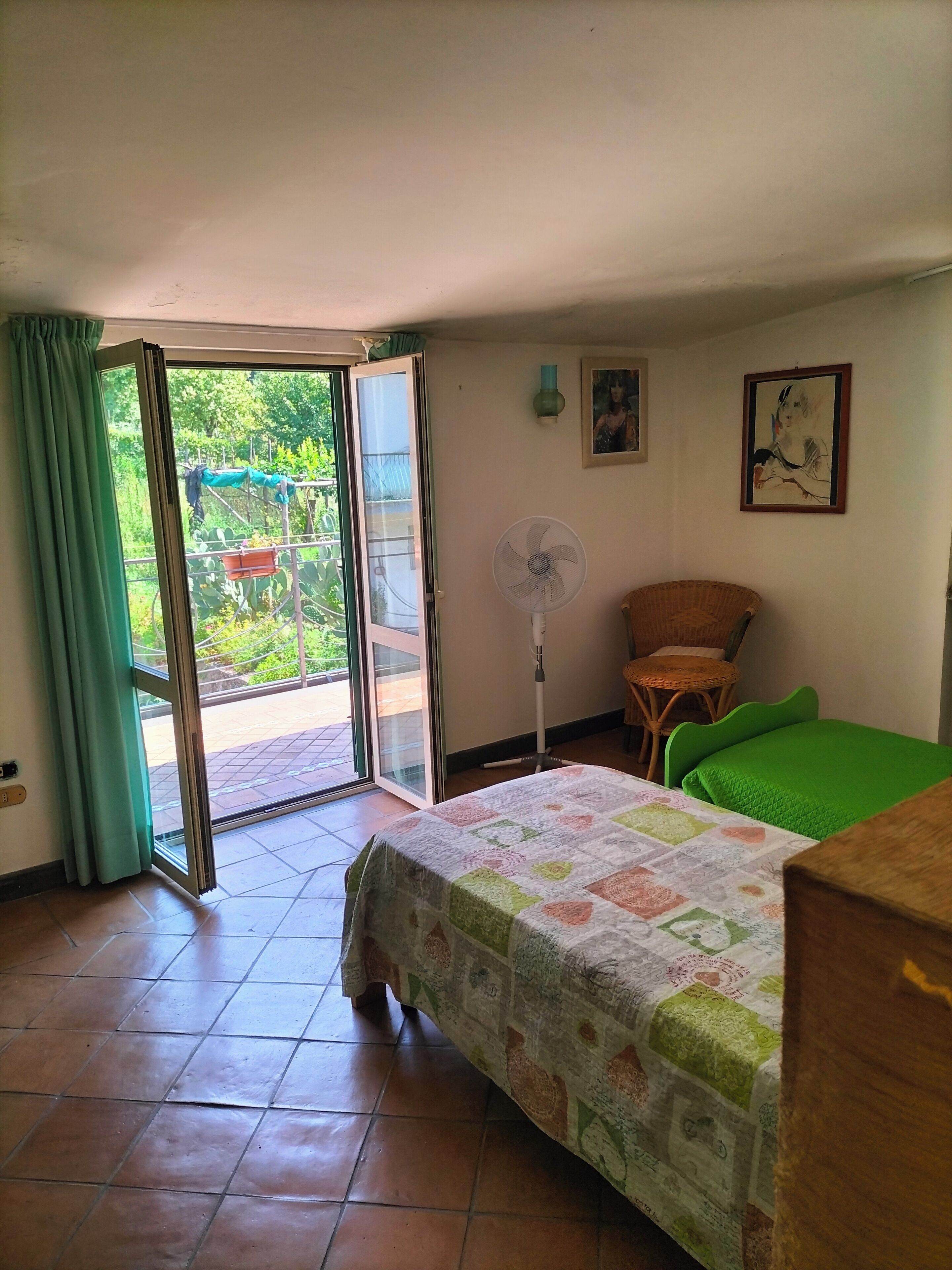A room with a bed, a chair, a fan, and a view through an open door to a garden.