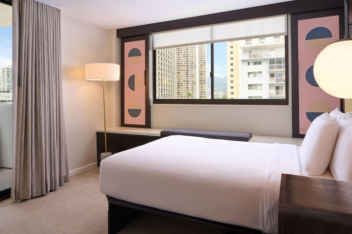 Habitación de hotel con cama, ventana con vistas a edificios, lámpara y cortina.