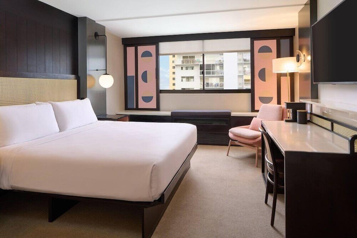 Une chambre d’hôtel avec un grand lit, un bureau, une chaise, une télévision et une fenêtre donnant sur des immeubles.