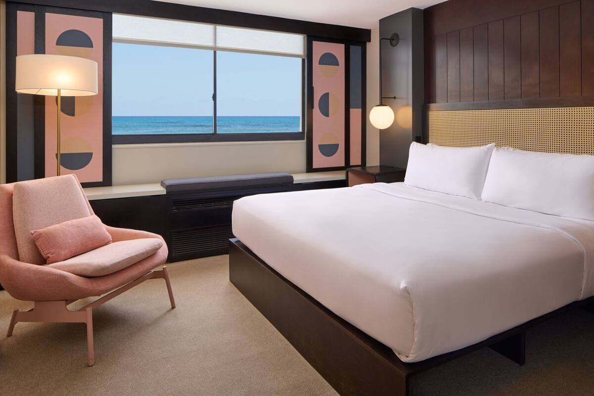 Une chambre d’hôtel avec un lit, une chaise, une grande fenêtre donnant sur la mer et des lampes fixées au mur.