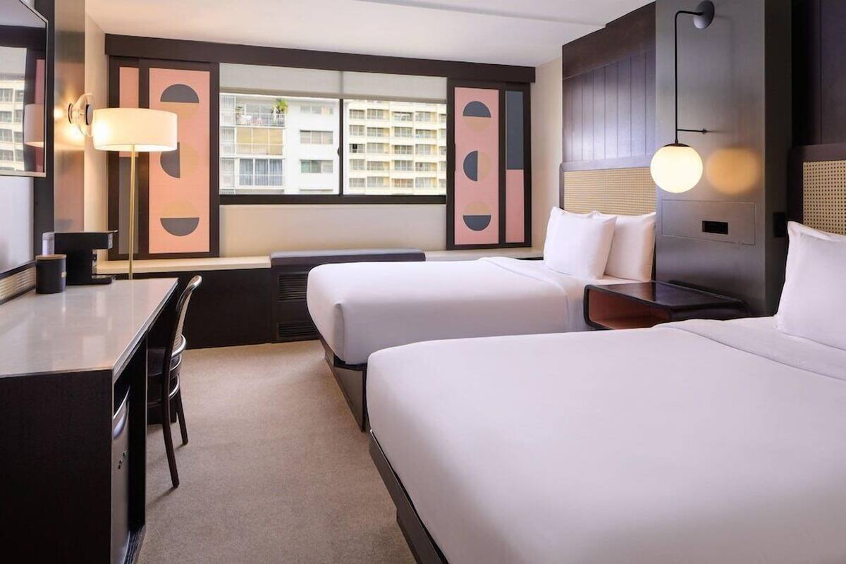 Une chambre d’hôtel avec deux lits, un bureau, une lampe et une vue sur les bâtiments par la fenêtre.