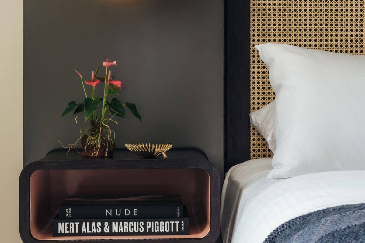 Une chambre d’hôtel moderne avec un lit, une table de chevet ornée d’une plante, un livre intitulé « NU », et un luminaire suspendu.