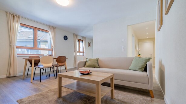 A living room with a sofa, a coffee table, and a dining area with chairs and a table. - Leaf Apartment in Zurich Oerlikon (Zürich)