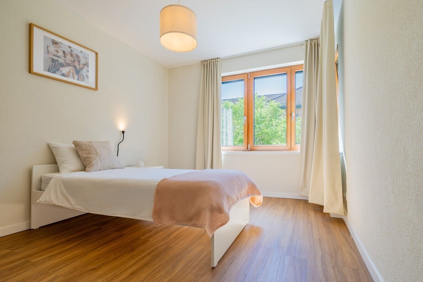 A bedroom with a bed, a bedside lamp, a window with curtains, a framed picture on the wall, and wooden flooring.