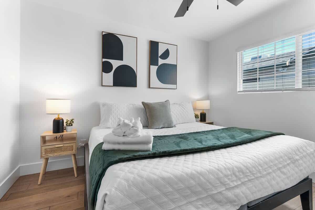 A modern bedroom with a large bed, two bedside tables with lamps, abstract wall art, and a window with blinds.