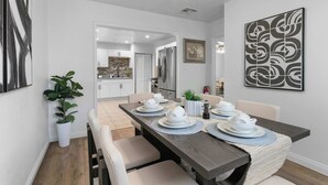 Un coin repas avec une table en bois dressée pour quatre personnes, entourée de chaises. Un pot de plante se trouve à gauche et une œuvre d’art encadrée est accrochée au mur, à droite.