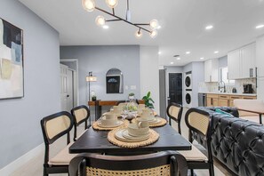 A modern kitchen with white cabinets, a dining area with a dark table and woven placemats, and a black leather sofa. - Grand 8BR Oasis – Pool, Spa, Arcade Games (North Las Vegas)