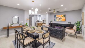 A modern dining area with a black dining table, beige cushioned chairs, and a chandelier. - Grand 8BR Oasis – Pool, Spa, Arcade Games (North Las Vegas)