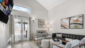 Uma sala de estar moderna com um sofá de couro preto, poltronas cinzas e uma mesa de centro. Há uma TV de tela plana na parede, um abajur e obras de arte nas paredes.
