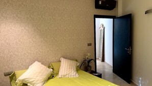 Ein Schlafzimmer mit Bett, Kissen, einem Nachttisch, einer Topfpflanze und einer Tür zu einem Flur.