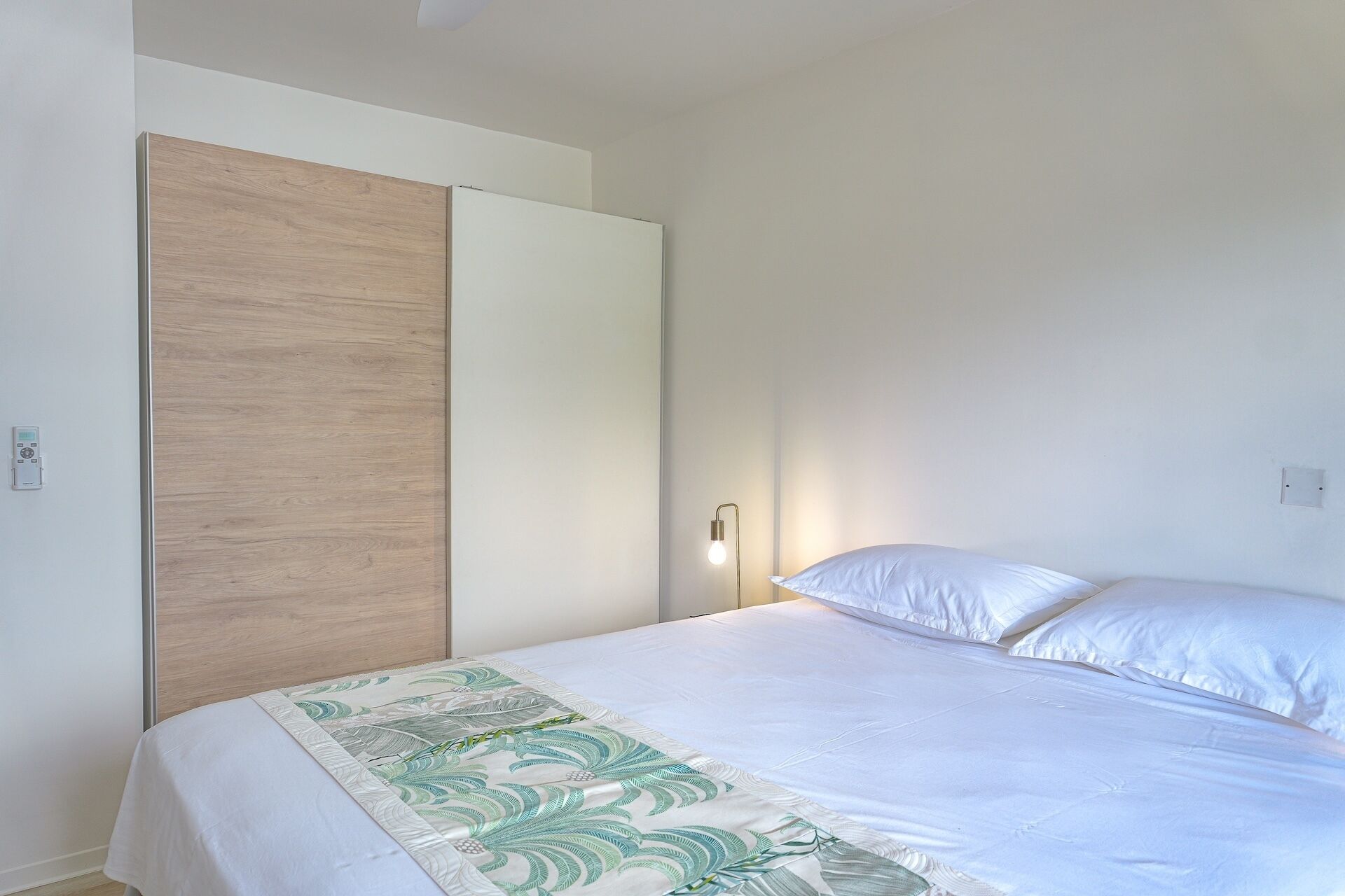 Un dormitorio con cama, ropa de cama blanca, cabecera de madera, mesita de noche y lámpara fijada a la pared.