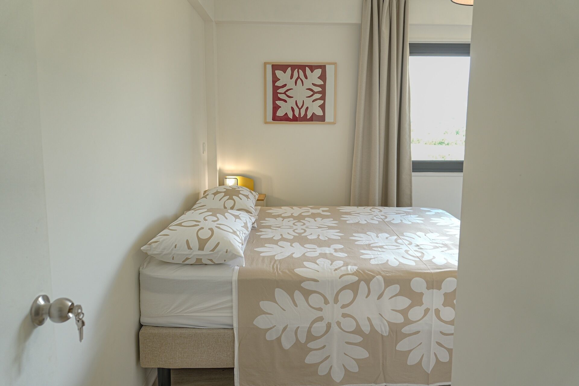 A bedroom with a bed, a bedside table with a lamp, a window with curtains, and a wall-mounted artwork.