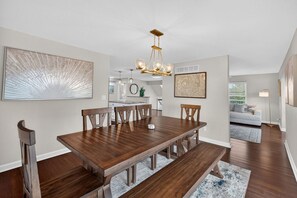 A dining area with a wooden table, benches, and a chandelier. There is a kitchen in the background with a white countertop and a hanging light fixture. - Luxe Sandbridge Beach, Waterfront, Pool, Arcade (Virginia Beach)