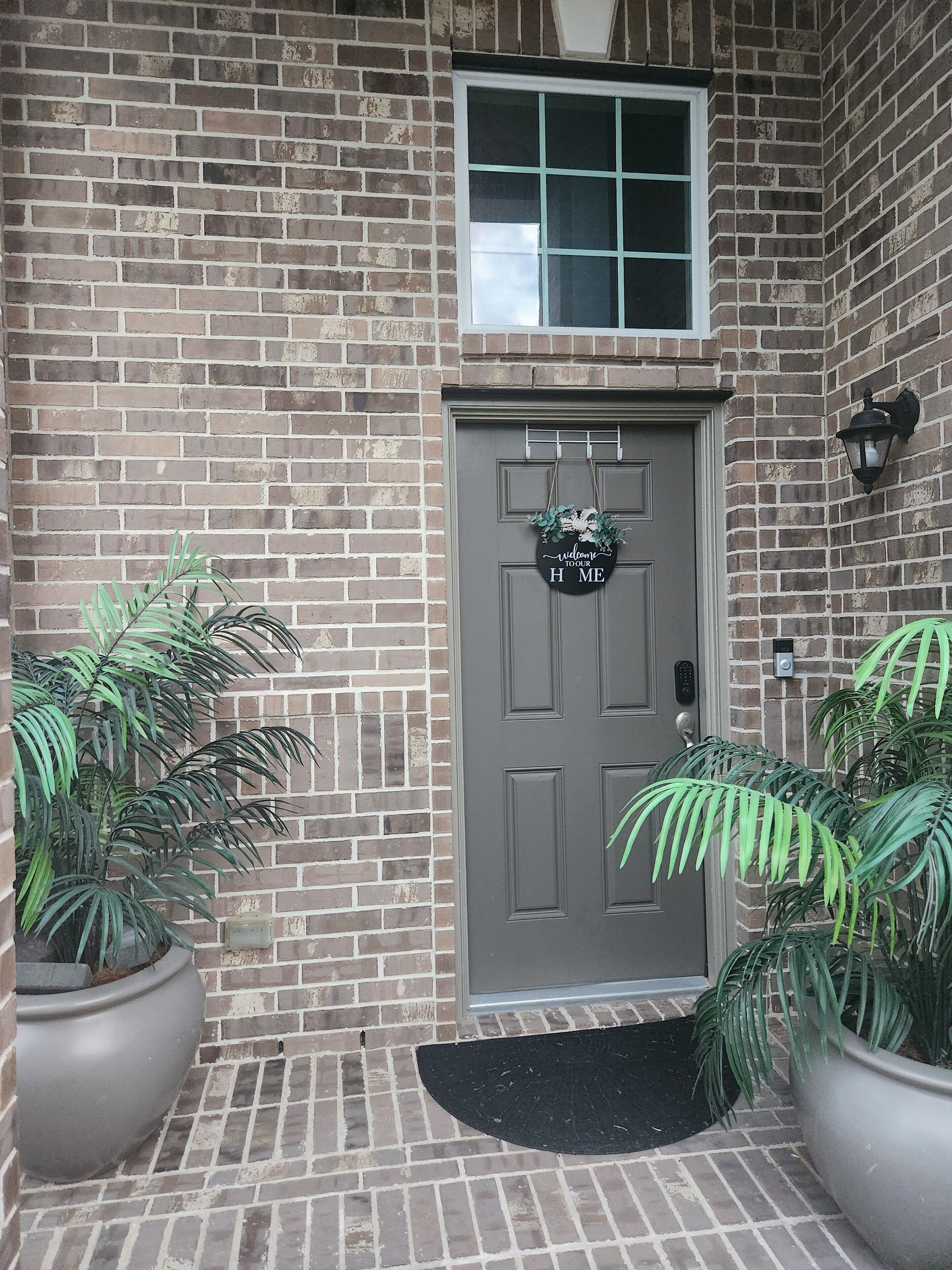 A brick building with a grey door, a welcome sign, and two potted plants.