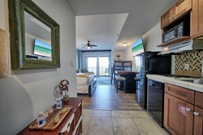 A modern kitchen with a dishwasher, microwave, and wooden cabinets. There is a dining area with a table and chairs, and a bedroom with a bed and window. - Rooftop Pool Hot Tub Arcade Coffee Shop and Great Location! (Panama City Beach)