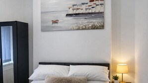 Una camera da letto ordinata con un letto, un comodino con una lampada e un quadro raffigurante una scena costiera.