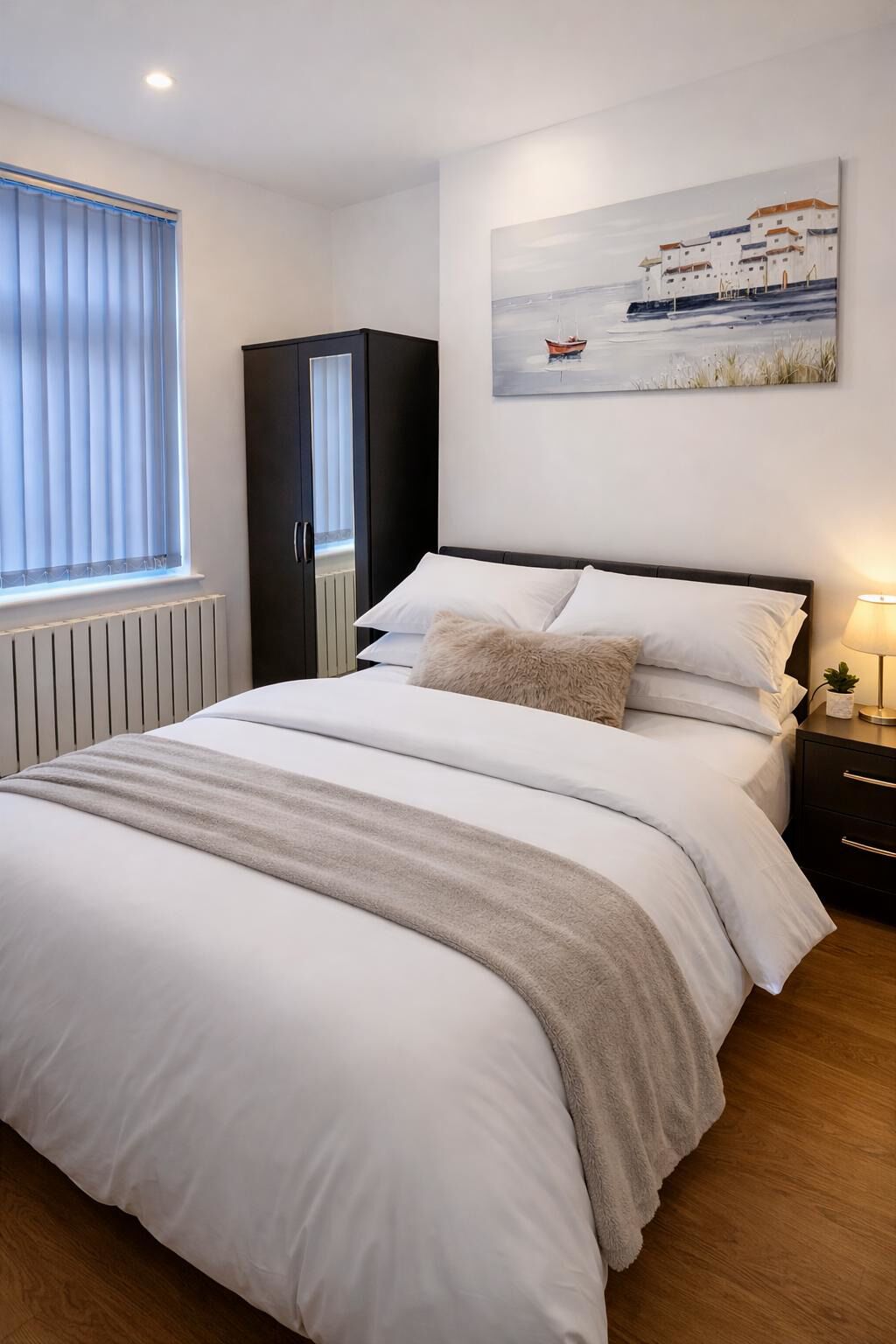 A bedroom with a large bed, a wardrobe, a radiator, a window with blinds, a painting of a boat, and a bedside lamp.