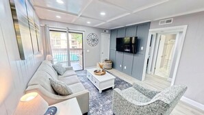 A modern living room with a grey sofa, a white coffee table, and a grey armchair. There is a flat-screen TV mounted on the wall and a window with a view of a balcony. - FANTASTIC FIND! Great deals and in the heart of everything! Beaches, golf, shops (Hilton Head Island)