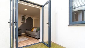 An open sliding glass door leading to a living room with a grey sofa, a patterned rug, and a framed picture on the wall. - Luxury 2BR Apartment | Balcony | Free Parking | 5min to Docks | Sleeps 6 (Gloucester)