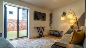 A modern living room with a sofa, a coffee table, a TV, and a balcony with a brick wall. - Luxury 2BR Apartment | Balcony | Free Parking | 5min to Docks | Sleeps 6 (Gloucester)