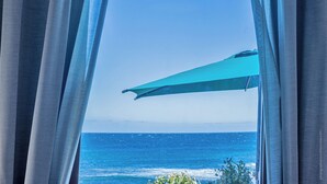 Door gordijnen is het uitzicht op de oceaan te zien, met een heldere lucht en een blauwe parasol.