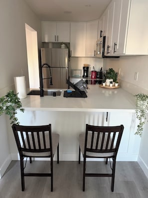 A modern kitchen with white cabinets, a stainless steel refrigerator, a sink, and a countertop with a coffee maker, toaster, and potted plants. There are two wooden chairs at the island.