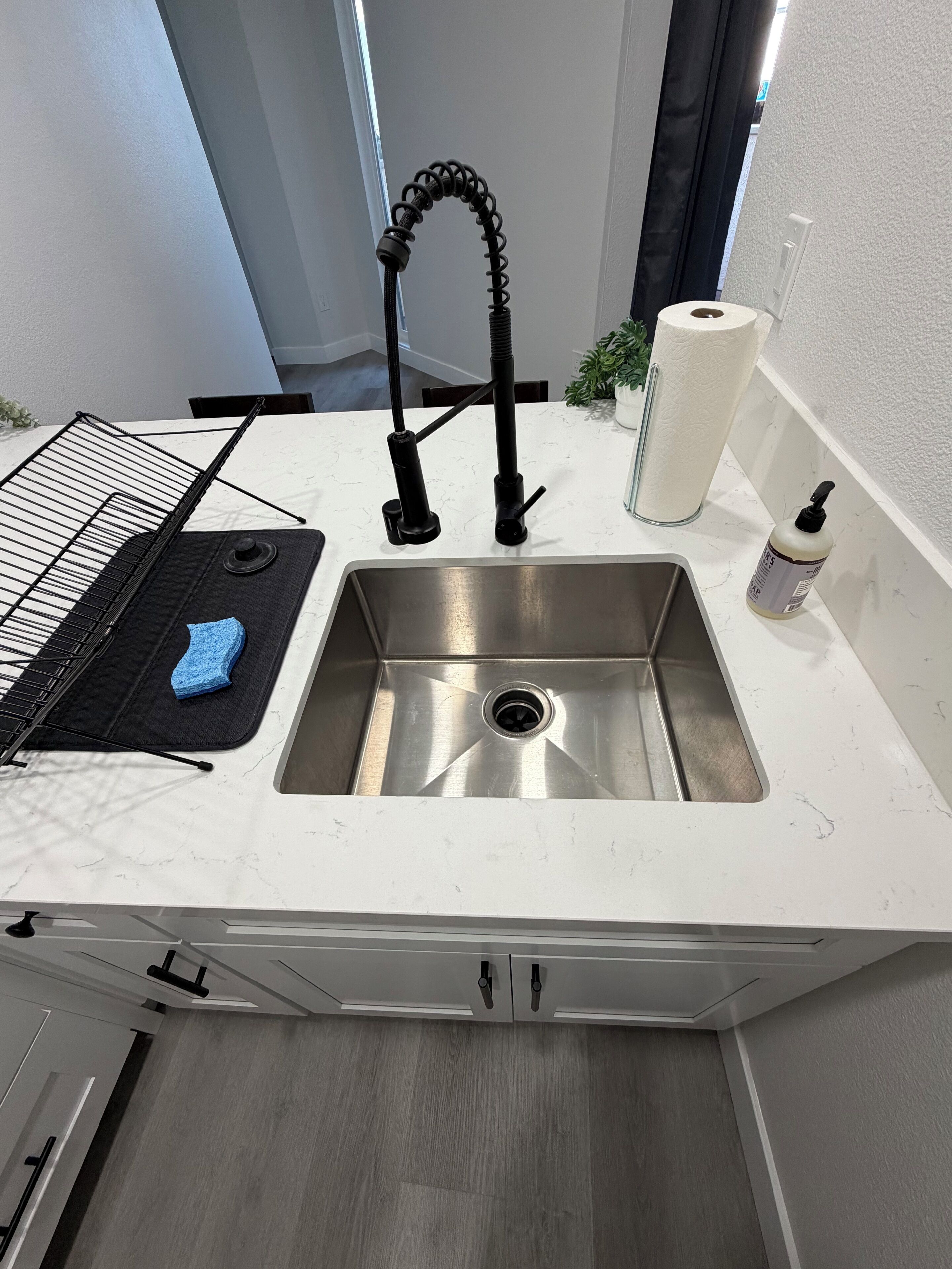 Keittiössä on ruostumattomasta teräksestä valmistettu pesuallas, metallinen pyykkikori, sininen pesusieni, pullo käsisaippuaa ja rulla paperipyyhkeitä.