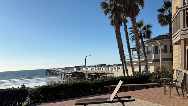 Eine Strandterrasse mit Liegestühlen, ein Steg im Hintergrund und Palmen.