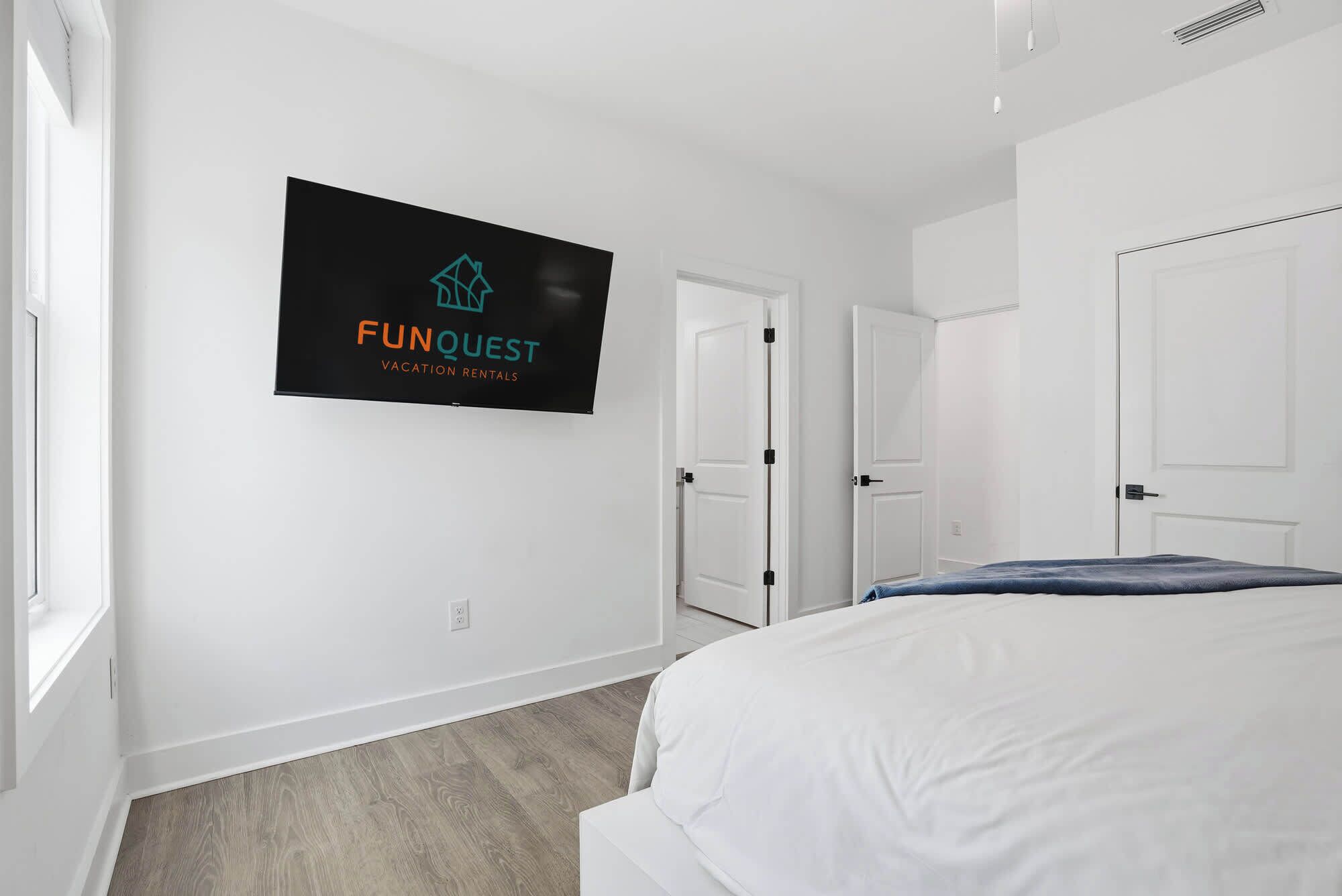 A modern bedroom with a bed, a TV mounted on the wall displaying 'FUNQUEST Vacation Rentals', and two white doors.