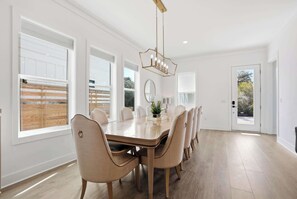 Une salle à manger avec une grande table en bois, des chaises de couleur beige, un lustre et de grandes fenêtres équipées de stores.