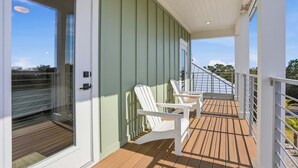 Un espacio al aire libre techado con una terraza de madera, sillas Adirondack blancas y un ventanal que da a una habitación.