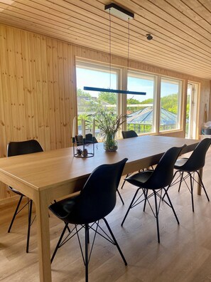 A modern dining area with a long wooden table, black chairs, and a view of greenery through large windows. - 2-family activity cottage (Rønde)