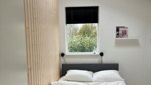A bedroom with a bed, a window with a view of greenery, a shelf with a magazine, and a wooden panel wall. - 2-family activity cottage (Rønde)