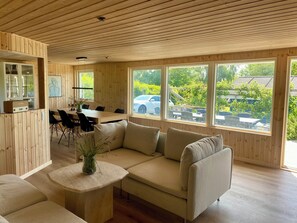 A wooden interior with a sofa, a dining table, and chairs, large windows with a view of the outside. - 2-family activity cottage (Rønde)
