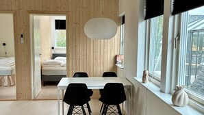 A room with a wooden ceiling, a dining table with chairs, a large window with blinds, and a bed in the adjacent room. - 2-family activity cottage (Rønde)