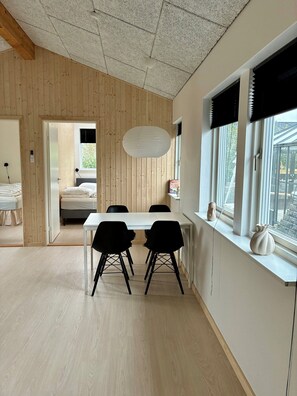 A room with a wooden ceiling, a dining table with chairs, a large window with blinds, and a bed in the adjacent room. - 2-family activity cottage (Rønde)