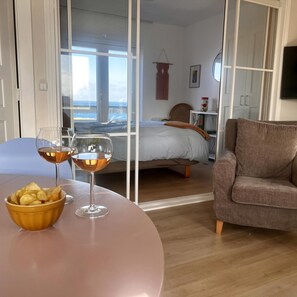 Une pièce comprenant un lit, un canapé, une table à manger avec un bol de chips et des verres à vin, et offrant une vue sur la mer grâce à de grandes fenêtres.