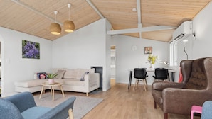 A modern living room with a sofa, armchairs, a dining table with chairs, a wooden ceiling, and a wall-mounted air conditioner.