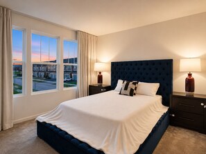 Une chambre à coucher avec un grand lit, deux tables de chevet, une fenêtre avec des rideaux et une vue sur un quartier résidentiel.