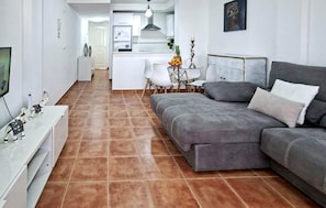 Un apartamento moderno con un sofá gris, una pequeña mesa de comedor con sillas, una zona de cocina y un televisor sobre un mueble blanco.