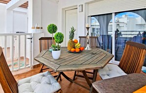 Una mesa de madera con un frutero, dos plantas en macetas y una vela en un balcón con sillas y cojines de madera.