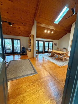 A spacious room with wooden floors, a dining table with chairs, a bookshelf, and a large window.