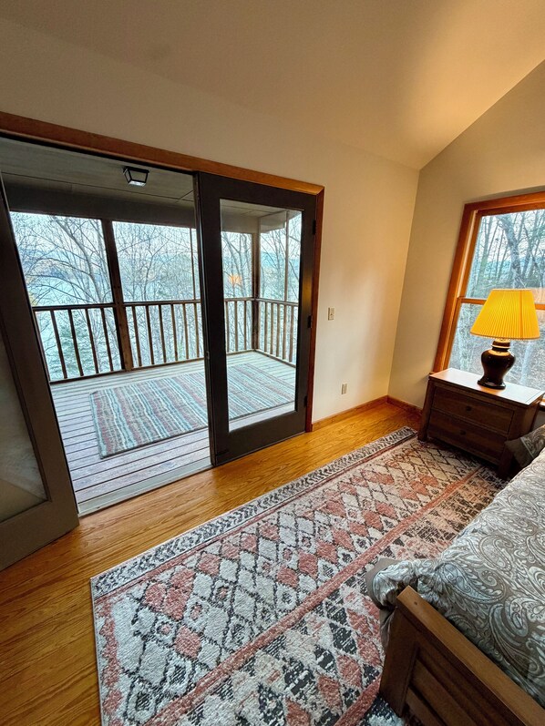 A bedroom with a large sliding glass door leading to a balcony, a wooden floor, a patterned rug, a bedside table with a lamp, and a bed with a patterned bedspread.